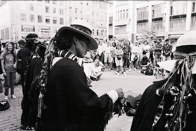 Crowded urban street scene featuring Indigenous performers in traditional regalia, likely during a cultural festival or prote...