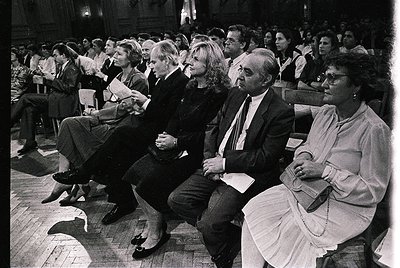 A black-and-white photo of an attentive audience in a formal indoor setting, likely a lecture or ceremony. Wooden pews and a ...