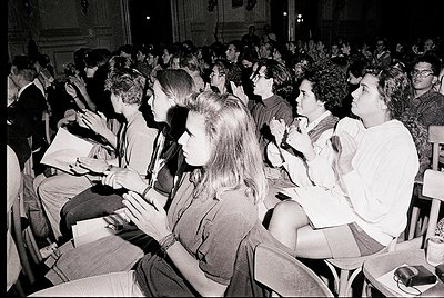 Crowded indoor lecture hall with seated students, likely 1960s–1970s. Wooden benches, formal attire (blazers, skirts), and cl...