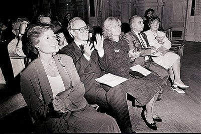 A black-and-white photo of an indoor audience in a formal setting, likely a 1970s conference or lecture. Six adults sit in wo...