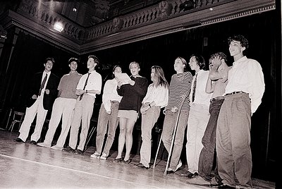Black-and-white stage performance featuring 9 actors in 1970s attire—men in dress shirts/ties, women in blouses/skirts or pan...