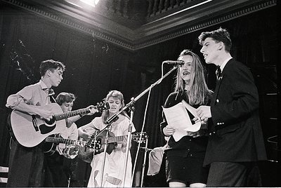 Vintage black-and-white photo of a folk/rock band performing live in a dimly lit indoor venue, likely 1960s–1970s. Four music...