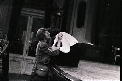 Two individuals embrace in an indoor setting, likely a theater or performance space. The person on the left wears a dark jack...