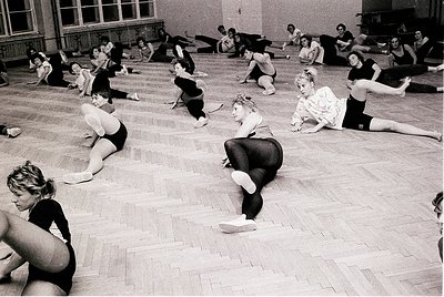 Vintage black-and-white photo of a ballet class in a large, wood-floored studio. Women in leotards and tights perform floor e...