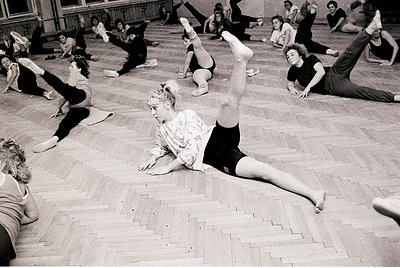 Group fitness class in a spacious indoor gym, likely mid-20th century. Women perform floor exercises in coordinated poses, em...