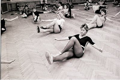 Group ballet class in mid-20th century studio, likely 1950s–1960s. Women in leotards and ballet slippers perform floor exerci...