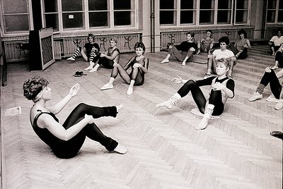 Black-and-white photo of a 1960s-70s ballet class in a large, wood-floored studio with high windows. Women in leotards and ti...