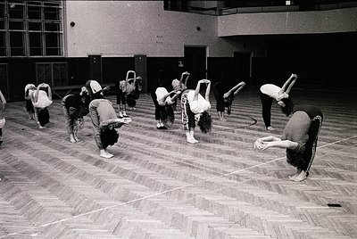 Group yoga/stretching session in an indoor gym, likely 1970s–1980s. Participants in loose clothing and headbands perform floo...