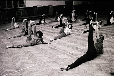 Group fitness class in a gymnasium, 1970s. Participants perform floor exercises in coordinated poses, likely inspired by Pila...