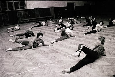 Group fitness class in a gymnasium, 1970s. Participants perform floor exercises in coordinated poses, likely part of a struct...