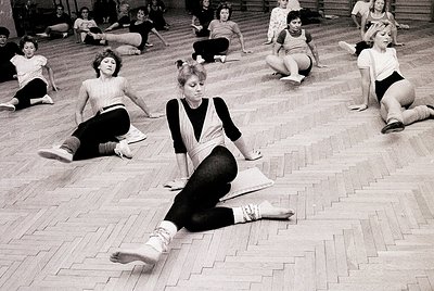 Group fitness class in a gymnasium, 1970s. Women in leotards and leg warmers stretch on wooden floors. Classic aerobic or yog...