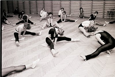 Group ballet class in a wooden-floored studio, 1960s–1970s. Women in leotards and tights perform floor exercises, emphasizing...