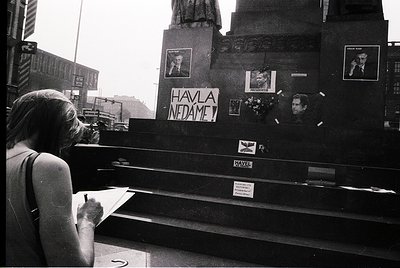 A woman in a sleeveless dress signs a memorial ledger at a public monument adorned with photos, flowers, and handwritten mess...