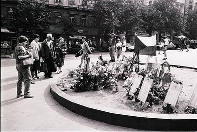 A solemn urban memorial scene from the late 20th century, likely Eastern Europe. A circular plaza features makeshift graves a...
