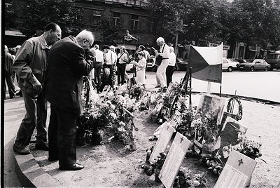 Black-and-white memorial scene with floral tributes and plaques arranged on a circular platform. Elderly men and women in 197...