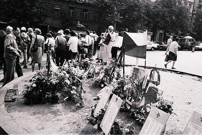 A solemn gathering at a memorial site, likely from the mid-20th century. Crowded urban street with people paying respects to ...
