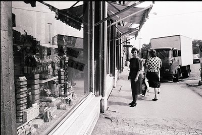 Vintage black-and-white street scene featuring a corner grocery store with shelves stocked in stacked cardboard boxes and jar...
