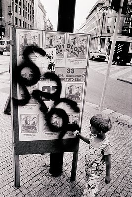 A child in a patterned sleeveless dress examines a vandalized public notice board in an urban setting, likely Eastern Europe....