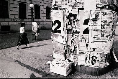 Vintage black-and-white street scene featuring a covered post office box overflowing with stacked flyers and ads. A woman in ...
