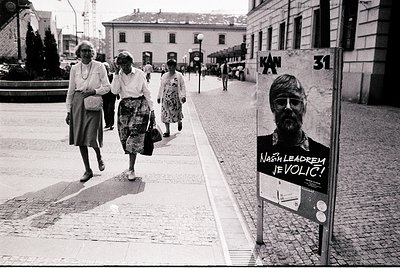 Black-and-white street scene featuring three women in 1970s-era dresses and headscarves crossing a cobblestone pedestrian cro...