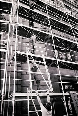 Mid-20th-century construction crew ascending metal scaffolding on a high-rise facade. Workers in casual attire (short-sleeved...