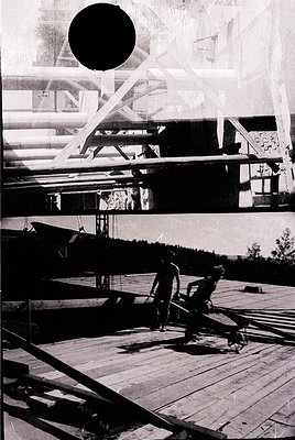 Vintage black-and-white photo of a wooden pier with two figures seated on benches, overlooking a forested shoreline. Structur...