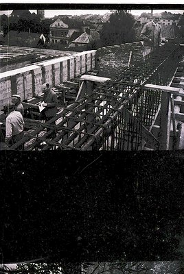 Mid-20th century construction site featuring reinforced concrete framing. Workers in casual attire (short sleeves, caps) asse...