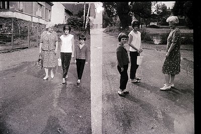 Two women and two children walking on a residential street, 1960s-1970s. The woman on the left wears a floral dress and carri...
