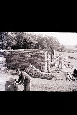 Mid-20th century bricklaying crew constructing a wall, likely . Workers in rolled-up sleeves, using bricks and mortar; stacke...