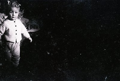 Black-and-white portrait of a young child in early 20th-century attire—buttoned sweater, trousers, and hat—posing indoors aga...