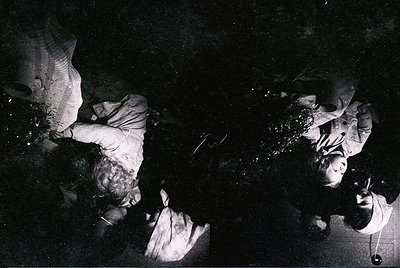 Black-and-white photo of two individuals engaged in textile work under dim lighting, likely a loom. Cloth appears partially w...