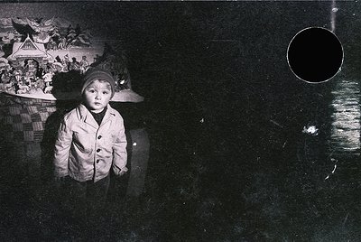 Black-and-white portrait of a young child in a heavy winter coat and knitted cap, standing indoors near a dark, reflective su...