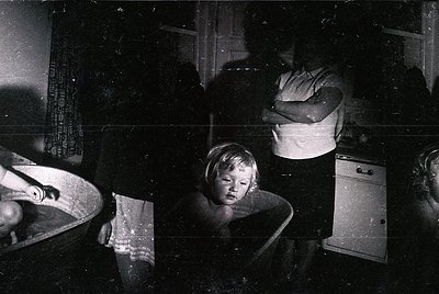 Vintage black-and-white photo capturing a dimly lit kitchen scene, likely mid-20th century. A woman kneels beside a child sea...