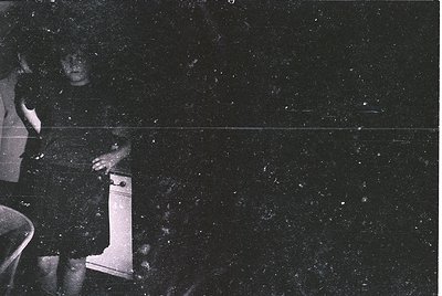 Vintage black-and-white photo of a person’s lower body seated on a wooden chair, holding a small, upright object resembling a...