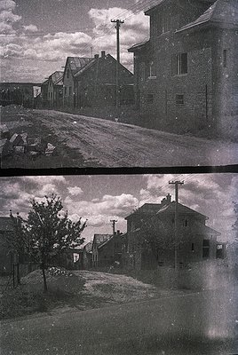 Mid-20th century urban street scene featuring brick buildings with exposed wiring and utility poles. Wet pavement suggests re...