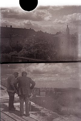 Two men in mid-20th-century suits stand on a rooftop overlooking a European cityscape, likely . Architectural details include...