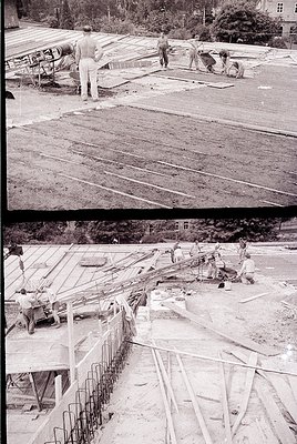 Mid-20th century construction site featuring concrete slab pouring and rebar reinforcement. Workers in utilitarian clothing l...