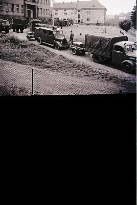 Vintage black-and-white photo of a rural roadside scene with vintage military trucks and a jeep, likely mid-20th century. Ope...