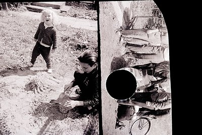 A candid 1970s-era black-and-white photo of a woman kneeling in a garden, tending to a small child standing beside her. The w...