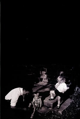 Black-and-white photograph of three children exploring a cemetery at dusk. Adult kneels beside a child in floral dress, while...
