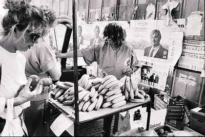 Street vendor stall showcasing bundled sausages and political campaign posters. Two women engage with products—one handling s...