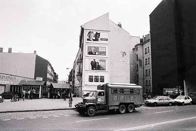 Urban street scene featuring a mid-20th-century European cityscape. Prominent white building with large posters of a cat and ...