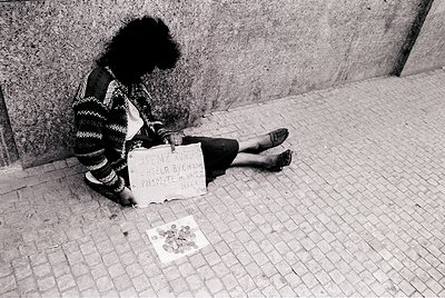 A person sits on a cobblestone street, leaning against a concrete wall, holding a handwritten sign reading *"I am a woman, he...