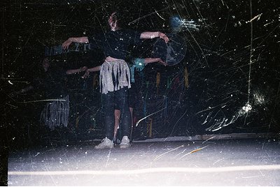 Two dancers in mid-motion against a shattered glass backdrop, likely a stage setting. The performer on left wears a sleeveles...