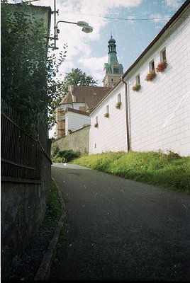 Historic European street scene featuring a narrow cobblestone path flanked by a whitewashed wall with hanging flower boxes. A...