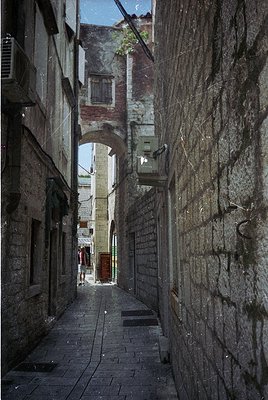 Narrow alleyway framed by weathered stone walls and arched passageways, likely Mediterranean. Cobblestone path leads to a dis...
