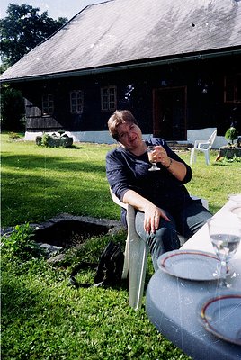 Relaxed individual in outdoor setting, holding a wineglass beside a rustic wooden house with white trim. Mid-century style fu...