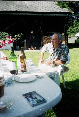 Vintage outdoor dining scene featuring a man in a floral-patterned shirt seated at a white-clothed table under a shaded canop...