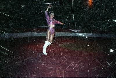 Circus performer mid-air on trapeze bar, dressed in 1970s-style leotard with geometric patterns and white boots. Dynamic shot...