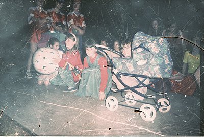 Vintage indoor gathering with children seated on floor, surrounded by luggage and a vintage stroller. Blurred, grainy quality...